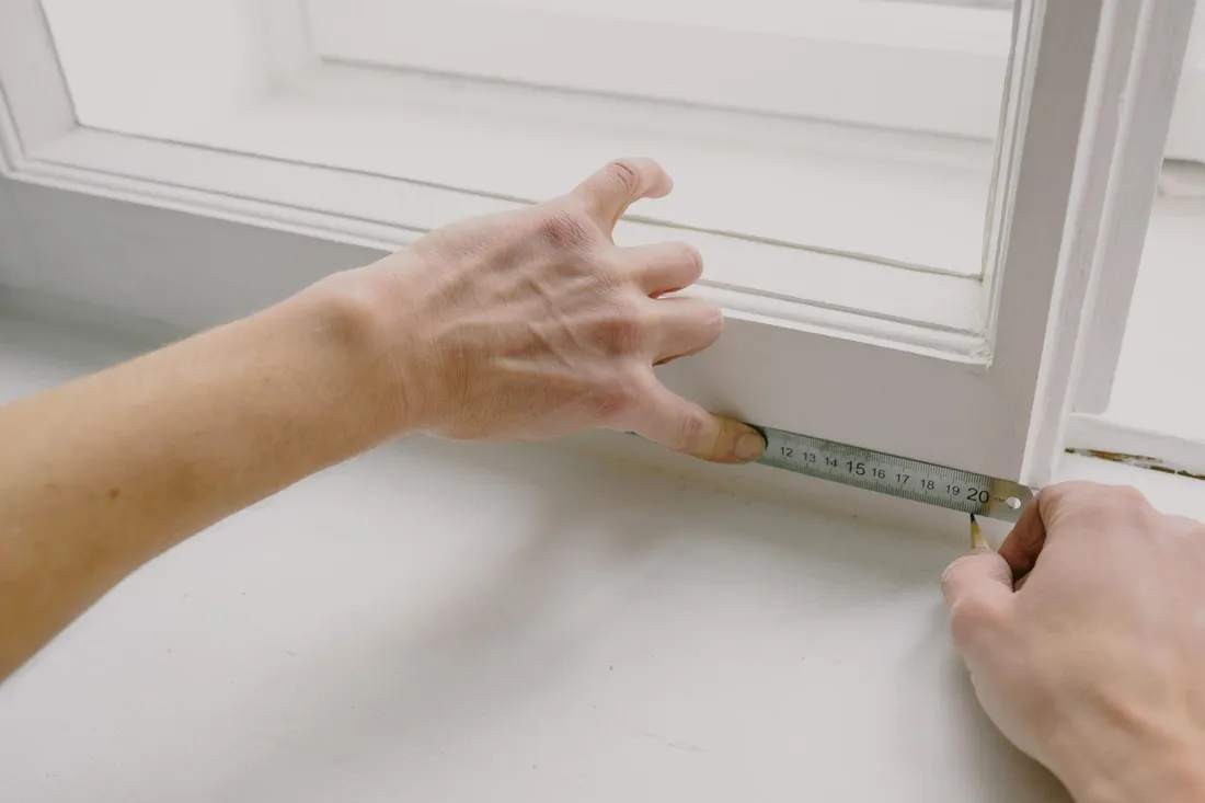 Zwei Hände messen mit einem Zollstock die Breite einer Fensterlaibung in einem hellen Raum.