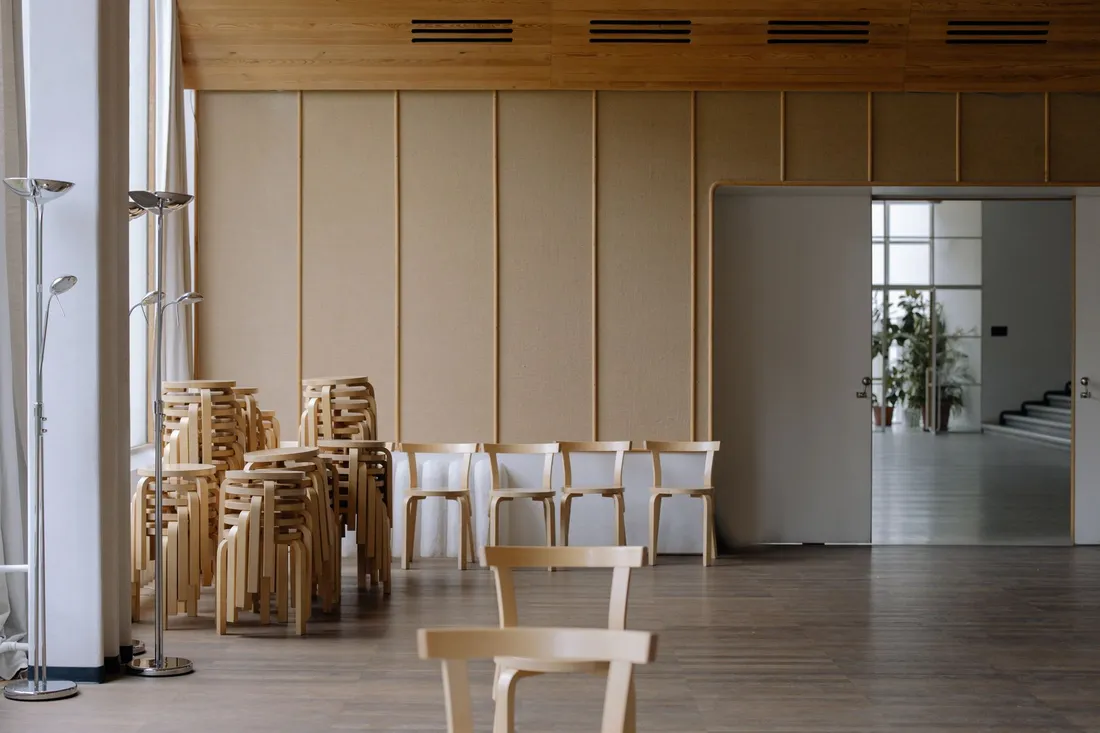 Gestapelte Holzstühle in einem leergeräumten Saal mit hohen Fenstern und offener Schiebetür.
