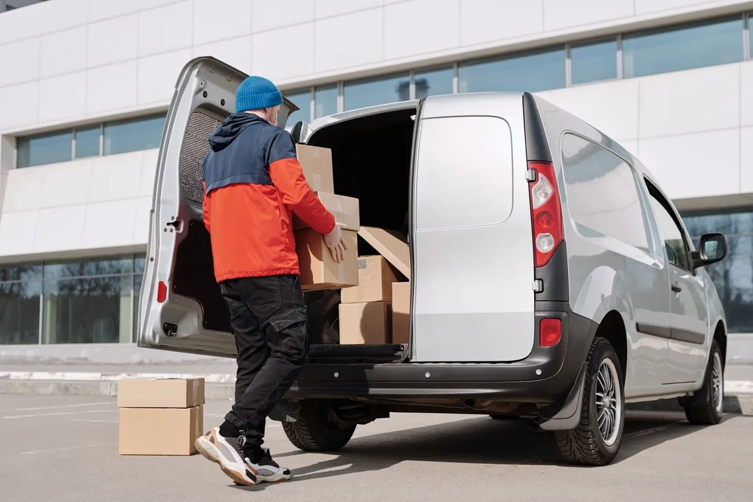 Ein Mann in Arbeitsjacke schiebt Kartons in den offenen Laderaum eines silbernen Transporters.