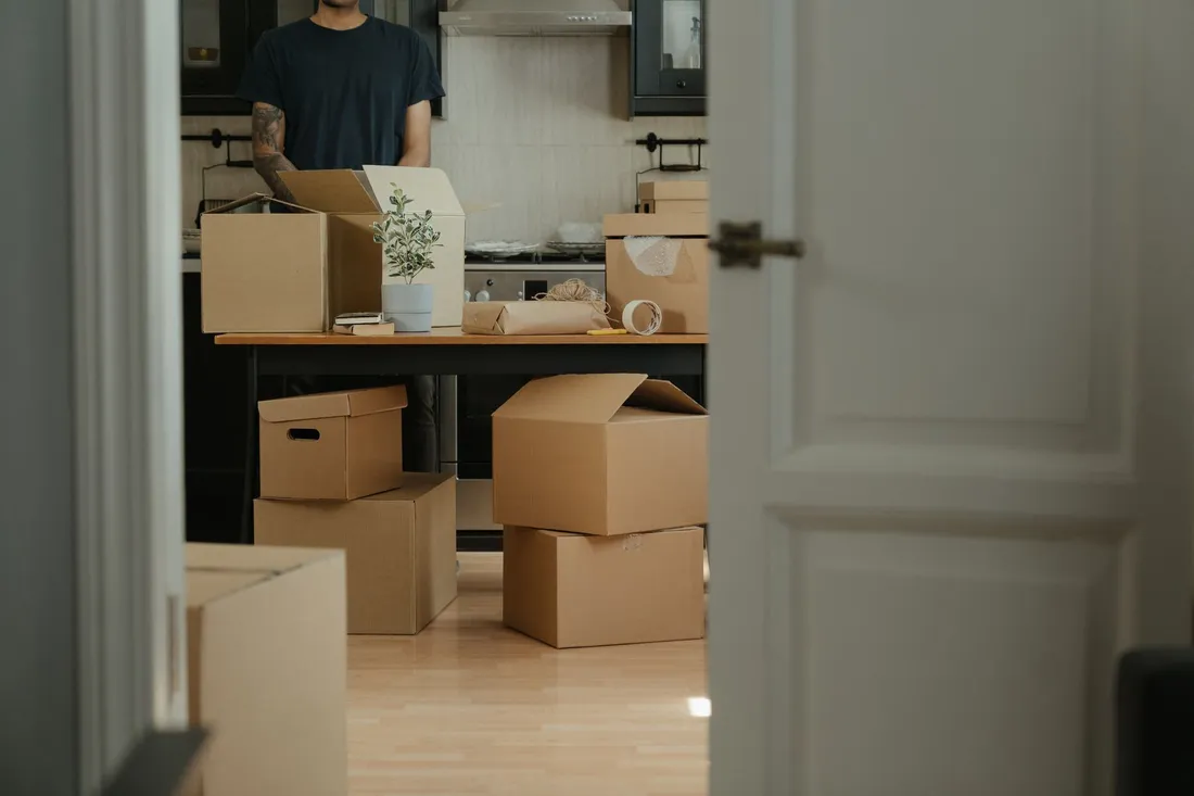 Halb gepackte Wohnung mit braunen Umzugskartons auf dem Boden und auf dem Tisch, im Hintergrund packt ein Mann.