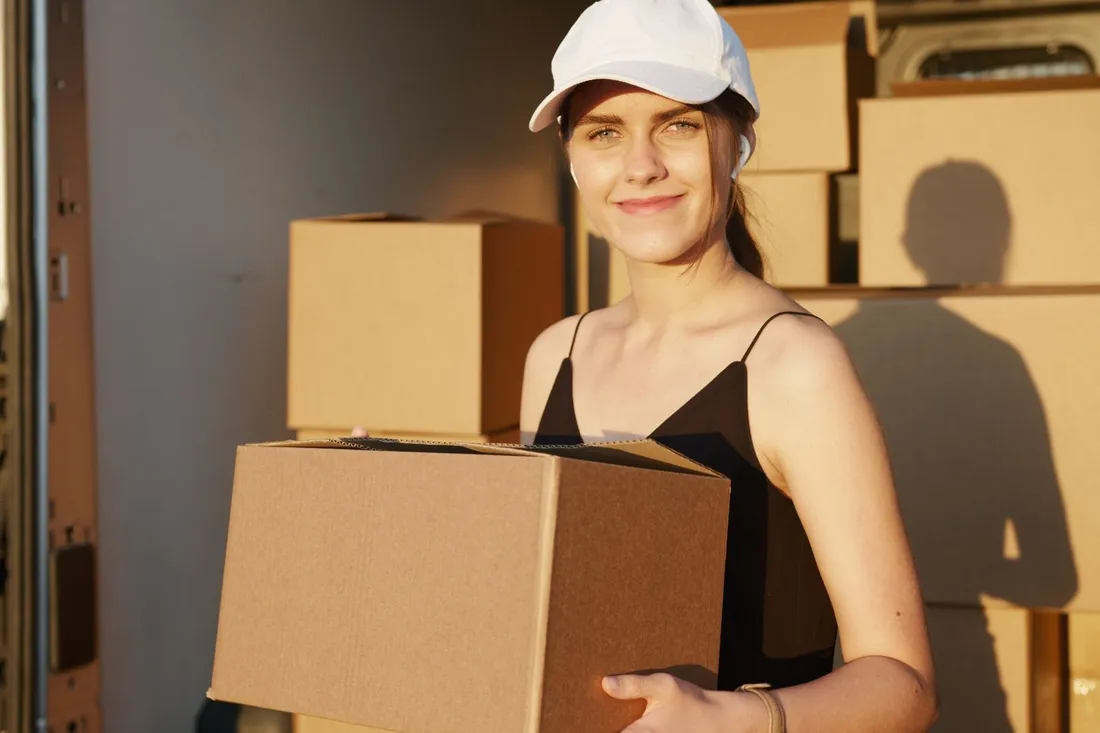 Junge Frau mit weißer Kappe trägt einen braunen Umzugskarton am geöffneten Laderaum eines Transporters