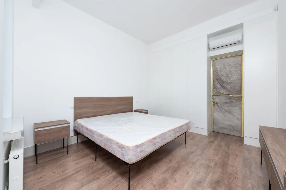 Leergeräumtes Zimmer mit nackter Matratze auf dem Bettgestell, zwei Nachttischen und Holzboden.