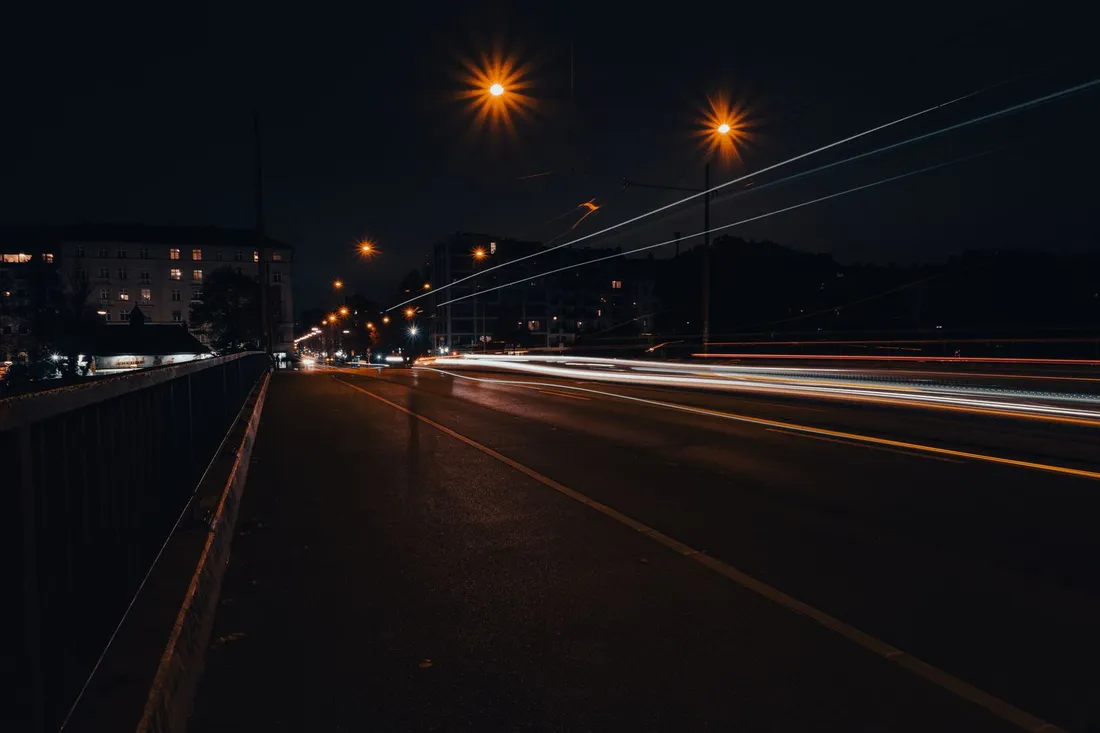 Nächtliche Straßenbrücke mit langen Lichtspuren vorbeifahrender Fahrzeuge unter orange leuchtenden Laternen.