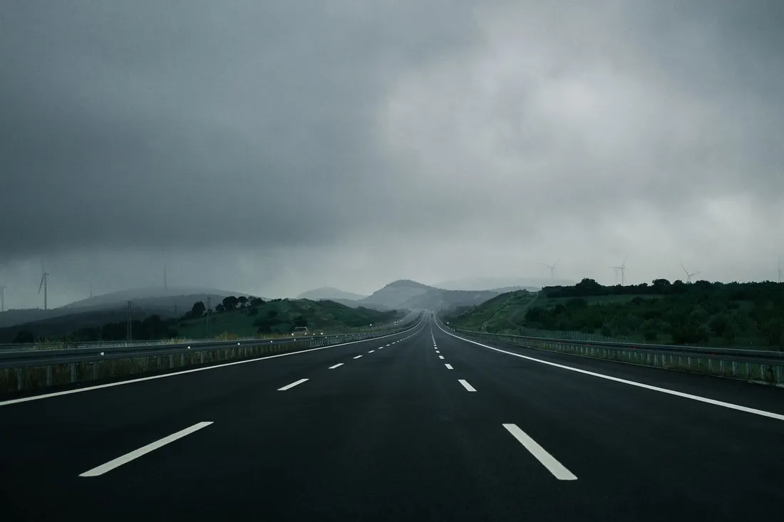 Dreispurige Autobahn unter dichten grauen Wolken, die leere Fahrbahn führt auf bewaldete Hügel zu.
