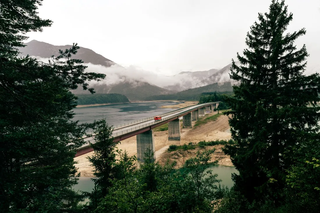 Lange Betonbrücke über einen Stausee, im Hintergrund bewaldete Berge in tief hängenden Wolken.