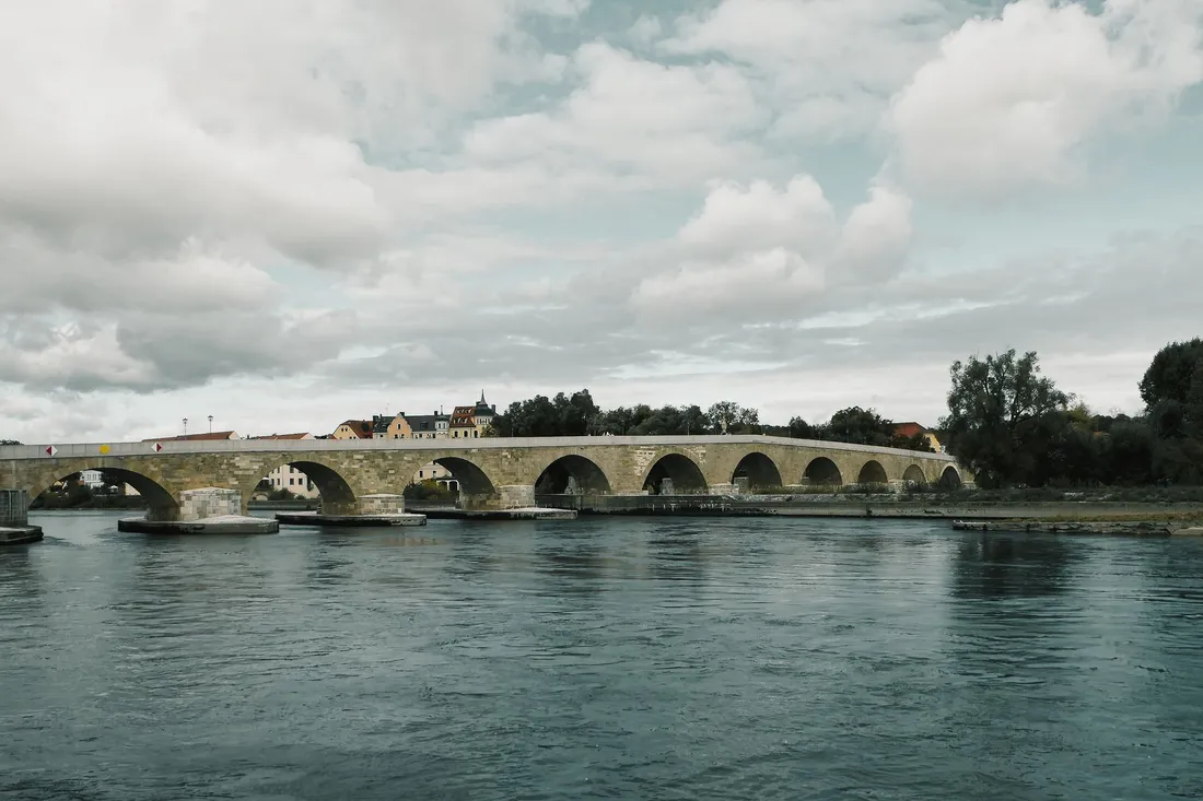 Alte steinerne Bogenbrücke spannt sich über einen breiten Fluss, dahinter Häuser und Bäume.