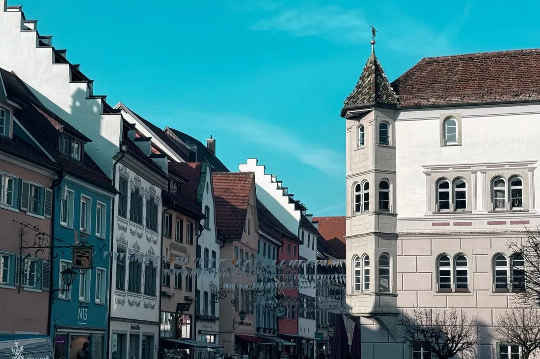 Sonnige Altstadtgasse mit bunten Giebelhäusern und einem Eckturm unter blauem Himmel.