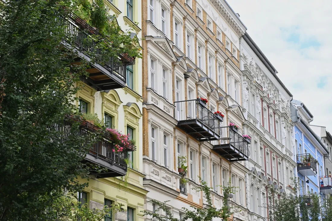 Hohe Altbaufassaden mit Stuck und schmiedeeisernen Balkonen voller Blumen in einer grünen Wohnstraße.